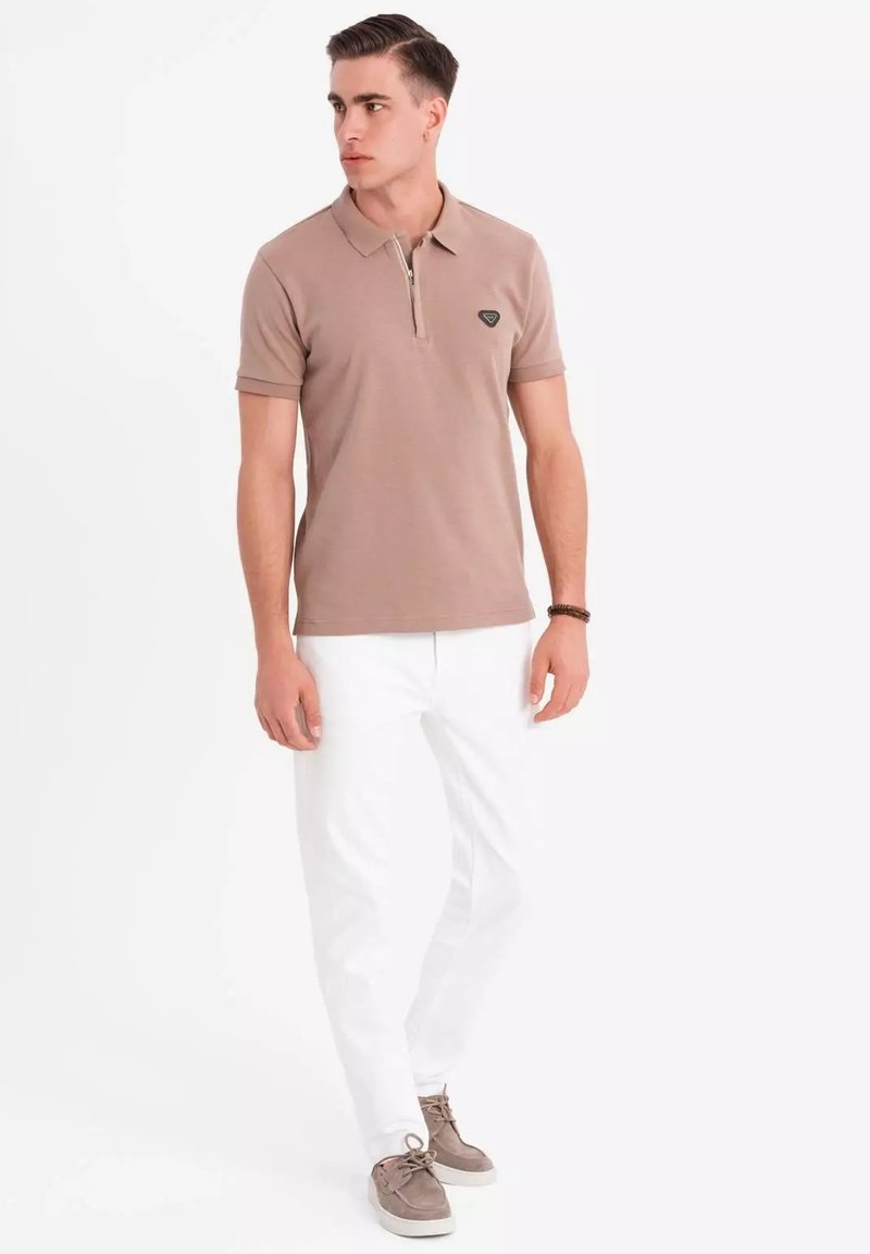 Polo de color beige con cremallera, de mangas cortas y un pequeño logo en el pecho. Combinado con pantalones blancos y zapatillas grises.