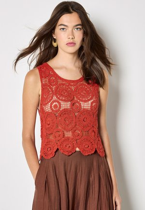 Junge Frau mit langen braunen Haaren trägt ein ärmelloses rotes Häkeloberteil und braune hoch taillierte Hosen, stehend vor einem schlichten Hintergrund.