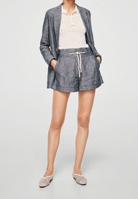 Žena v šedém lněném saku a odpovídajících šortkách na stažení, s bílým žebrovaným polo tričkem a béžovými tkanými polobotkami.