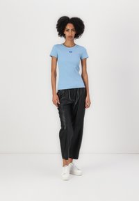 Camiseta ajustada de color azul claro con escote redondo y un pequeño logo, combinada con pantalones de cuero negros y zapatillas blancas.