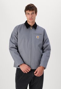 Veste de travail grise avec un col en velours côtelé noir, fermeture éclair sur le devant, et une poche poitrinère avec un patch logo. Le matériau semble durable et texturé.