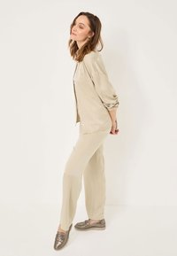 Beige, könnyű anyagú öltöny, laza szabással és gombos inggel. A modellen fémes loaferek vannak; sima textúra és minimális dizájn.