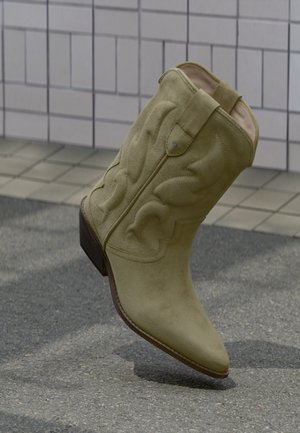 Botte de cowboy en daim couleur fauve avec coutures décoratives et talon en bois, positionnée en angle sur une surface pavée près d’un mur carrelé.