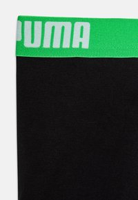 Črn material s zelenim pasom, na katerem je v belih črkah napisan "PUMA". Material je gladek, oblikovanje pa je osnovno, brez dodatnih vzorcev.