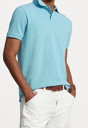 Homme portant un polo bleu clair à manches courtes avec col, pantalon blanc et ceinture tressée marron, mains dans les poches.