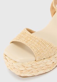 Sandalia de cuña con parte superior de piel beige y textura de paja trenzada. Diseño de punta abierta, con una plataforma gruesa para mayor altura.