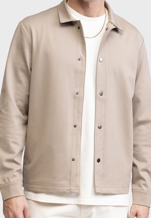 Veste beige à boutons portée sur une chemise blanche, associée à un collier chaîne doré et un pantalon blanc.
