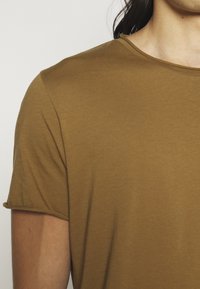 Gros plan d'une personne portant un t-shirt marron uni à manches courtes et col rond, montrant le haut du torse et le cou.