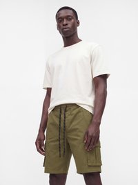 Homme portant une chemise blanche unie à manches courtes et un short cargo vert olive avec des cordons noirs sur un fond blanc uni.