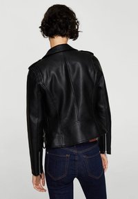 Henkilö, jolla on lyhyet kiharat hiukset, musta nahkatakki ja tummansiniset farkut, selkä kameraan päin yksinkertaisen taustan edessä.