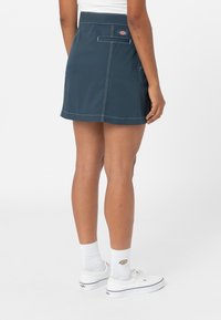 Námořnická modrá sukně ke kolenům s bílým prošitím, zadní kapsou a logem. Materiál má hladkou texturu a neformální design.