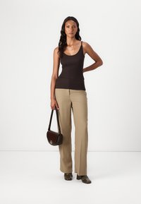 Un débardeur noir ajusté associé à un pantalon large beige, tenant un sac bandoulière marron, avec des chaussures sombres et à motifs. Fond blanc uni.