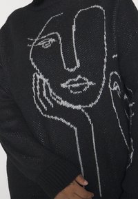 Pull en tricot noir avec un design en art linéaire blanc représentant un visage. Il a une coupe ample et des poignets côtelés pour une texture.