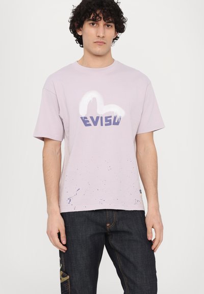 T-shirt lilas à manches courtes avec un logo graphique blanc et bleu, à coupe décontractée et à motif éclaboussé léger au bas.