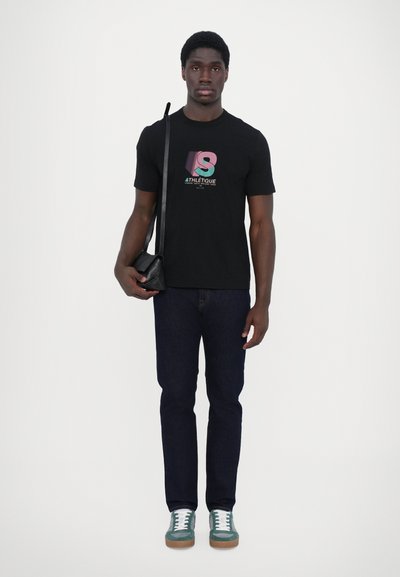 Camiseta negra con un gráfico colorido del logo "S", pantalones vaqueros azul oscuro y zapatillas verdes con cordones blancos, sosteniendo una pequeña bolsa negra de mano.