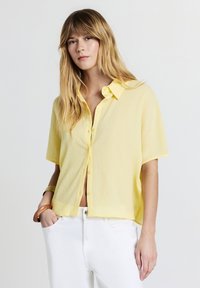 Giovane donna con lunghi capelli biondi che indossa una camicia gialla chiara a maniche corte con bottoni e pantaloni bianchi, in piedi con una mano nella tasca.