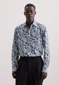 Camicia bianca con motivo floreale blu e verde, maniche lunghe e colletto classico, abbinata a pantaloni neri su misura.