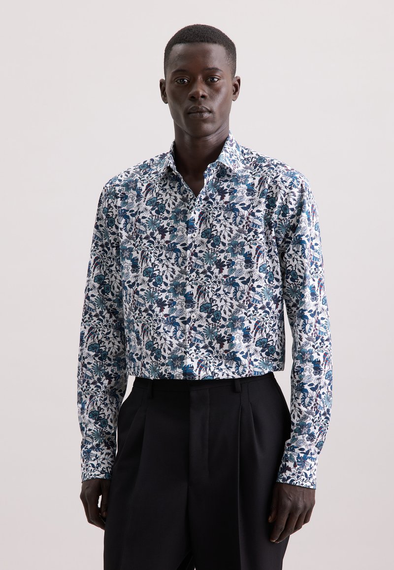 Camicia bianca con motivo floreale blu e verde, maniche lunghe e colletto classico, abbinata a pantaloni neri su misura.