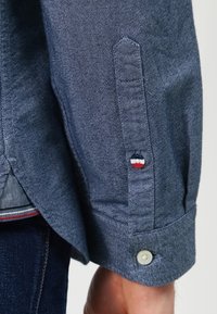 Knapp-up denimskjorta med subtil textur, mörkblå färg, kontrasterande vit knapp och en liten röd, vit och blå logo på ärmen.