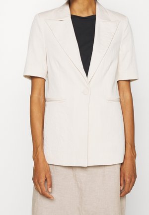Femme portant un blazer beige à manches courtes par-dessus un top noir, associé à un pantalon beige, vue du cou jusqu'à la mi-cuisse.