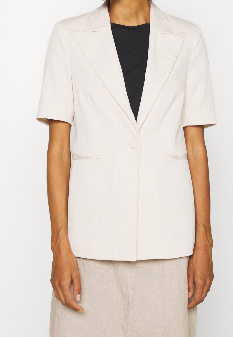 Femme portant un blazer beige à manches courtes par-dessus un top noir, associé à un pantalon beige, vue du cou jusqu'à la mi-cuisse.