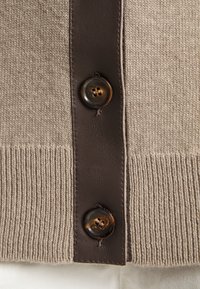 Béžový pletený cardigan s tmavě hnědým koženým límcem a dvěma kulatými hnědými knoflíky s lesklým povrchem. Jemná ribbová textura.
