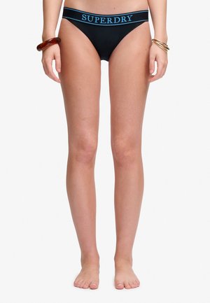 Widok z przodu osoby noszącej czarne majtki z niskim stanem z niebieskim paskiem z logo "SUPERDRY", stojącej boso z bransoletkami na obu nadgarstkach.