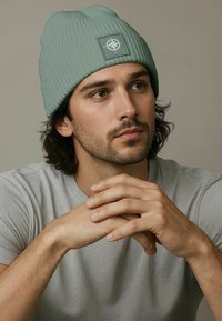 Light green ribbed beanie with a logo patch, worn by a person, paired with a gray t-shirt, featuring long dark hair and casual posture.