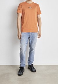 Oranžové bavlněné tričko s kulatým výstřihem a logem Calvin Klein, spojené se světle modrými džíny a černými teniskami s vzorem.