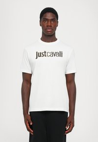 Balta kokvilnas T-kreklā ar melnu un zelta "Just Cavalli" logo priekšpusē. Īsas piedurknes un apaļa kakla izgriezuma forma.