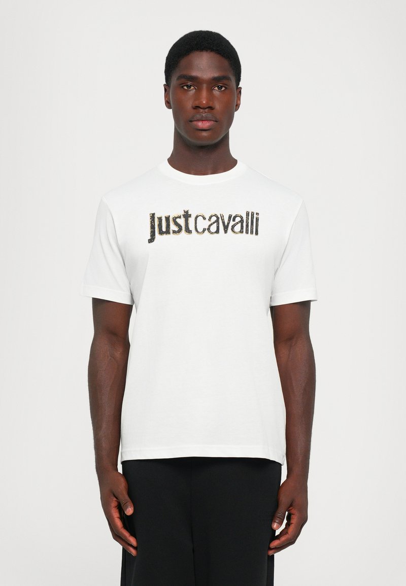 Balta kokvilnas T-kreklā ar melnu un zelta "Just Cavalli" logo priekšpusē. Īsas piedurknes un apaļa kakla izgriezuma forma.