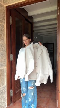 Blazer bianco over size con due bottoni e tasche frontali, appeso a una gruccia. Indossato con jeans in denim blu caratterizzati da motivi floreali e strappi.
