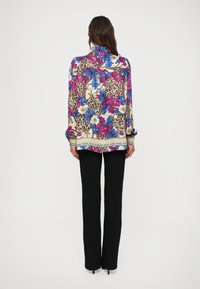 Kvinde iført en langærmet bluse med blomster- og dyreprint samt sorte lige bukser, stående med ryggen til på en ensfarvet baggrund.