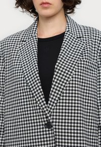 Blazer a quadretti bianchi e neri con chiusura a un bottone, rever a punta e taschino sul petto, abbinato a una maglia nera sotto.