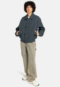 Person with curly hair wearing a dark blue buttoned jacket, light faded loose jeans, and white sneakers, standing with hands in pockets.