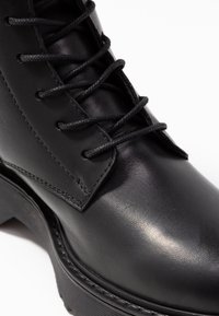 Botte en cuir noir avec une finition lisse, une couture détaillée et des lacets plats noirs, dotée d'une semelle épaisse et texturée.