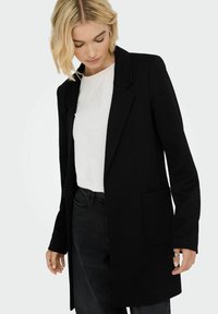 Kvinna med kort blont hår, iklädd en texturerad svart kavaj över en vit skjorta, tillsammans med svarta jeans mot en enkel bakgrund.