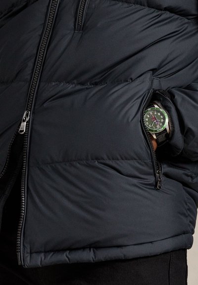 Čierna prešívaná bunda s čiastočne otvoreným zipsom na bočnom vrecku, ktoré odhaľuje náramkové hodinky so zeleno-čiernym ciferníkom na zápästí osoby.