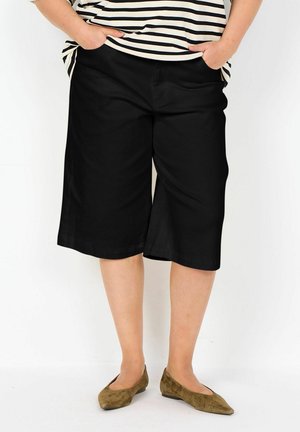 Person trägt schwarze knielange, weite Shorts, gestreiftes Hemd und braune spitze Ballerinas, mit den Händen in den Taschen vor einfarbigem Hintergrund.