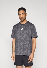 Γκρι t-shirt με μοτίβο tie-dye, με στρογγυλή λαιμόκοψη, κοντά μανίκια και λευκό λογότυπο στο κέντρο του στήθους. Φορεμένο με μαύρο αθλητικό σορτς.