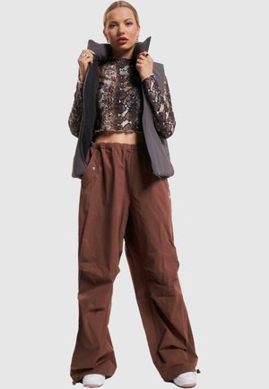 Grå vadderad väst över ett par bruna byxor med elastisk midja. Toppens långärmade cropp har ett reptilmönster, och vita sneakers fulländar outfiten.