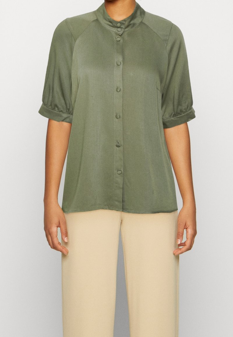 Femme portant un chemisier vert olive boutonné avec des manches courtes bouffantes et un pantalon droit beige, debout devant un fond uni.