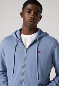 Lichtblauwe zip-up hoodie gemaakt van zachte stof, met een voorkant met rits, een capuchon met trekkoord en een klein rood Levi's-logo op de borst.