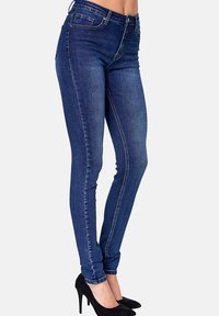 Jeans de denim azul ajustados con una cintura de media altura, diseño de cinco bolsillos y tejido elástico. Llevados con tacones altos negros para crear contraste.