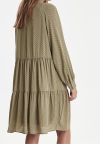 Khaki zelené šaty vyrobené z lehké tkaniny s černými puntíky, s vrstveným designem, dlouhými rukávy a manžetami na knoflíky.