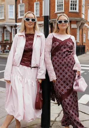 Zwei Frauen mit Sonnenbrillen und übereinander getragenen pinken und burgunderfarbenen Kleidern stehen an einem Straßenpfosten auf dem Bürgersteig einer Stadt.