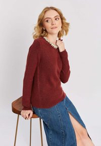 Sarkans vilnas džemperis ar apaļu kakla izgriezumu, ar teksturētu apdari un rotājošu kakla līniju. Zils džinsa svārks ar augstu šķēlumu un izšuvuma detaļām.