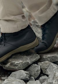 Czarne wodoodporne buty trekkingowe z teksturowaną skórzaną cholewką i brązową gumową podeszwą, z sznurowadłami i akcentującą pętlą w pobliżu kostki.