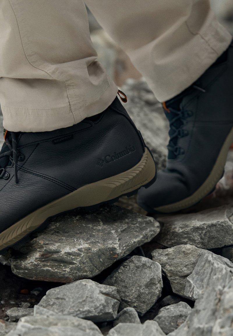 Botas de senderismo impermeables negras con un empeine de cuero texturizado y una suela de goma marrón, con cordones y un lazo decorativo cerca del tobillo.