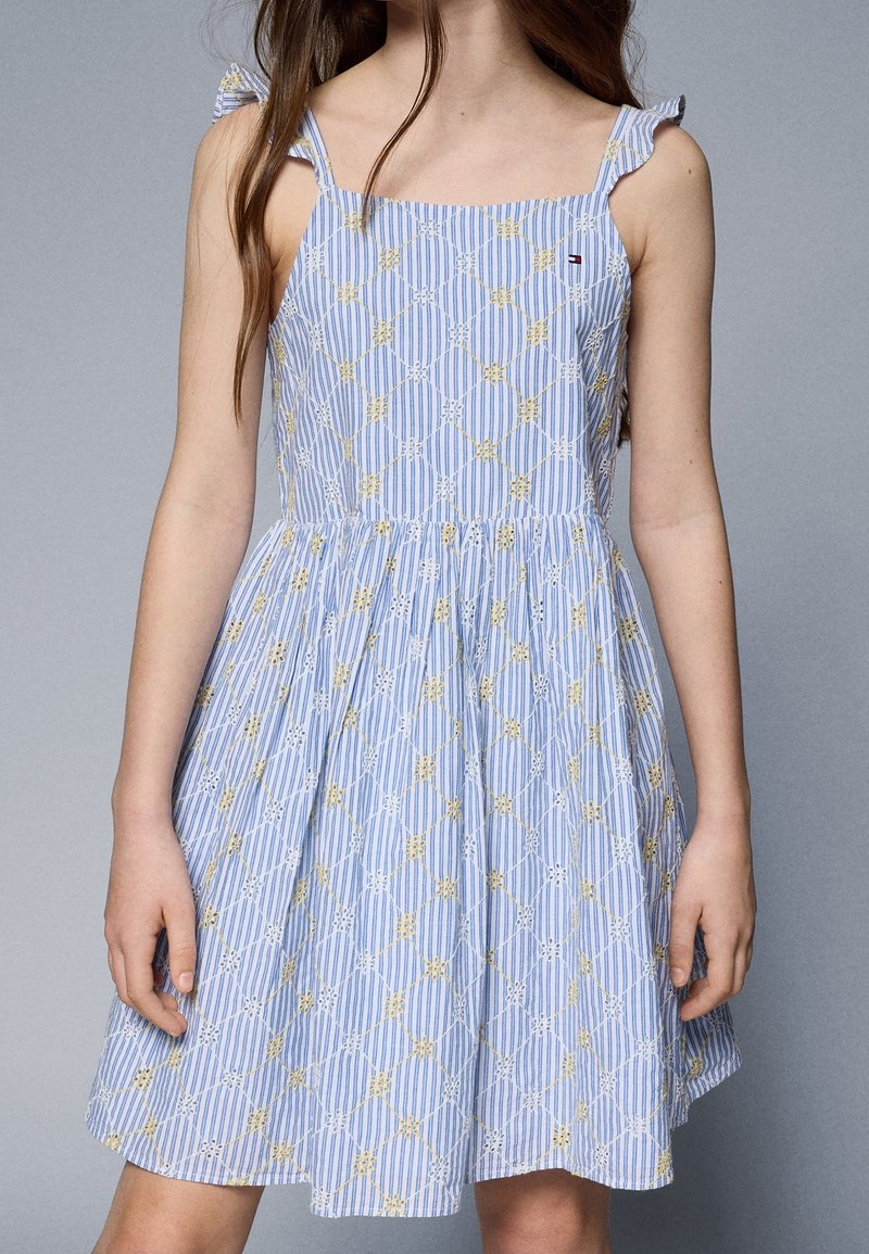 Fille portant une robe sans manches rayée bleu et blanc avec de petites broderies florales jaunes et blanches, debout devant un fond gris uni.